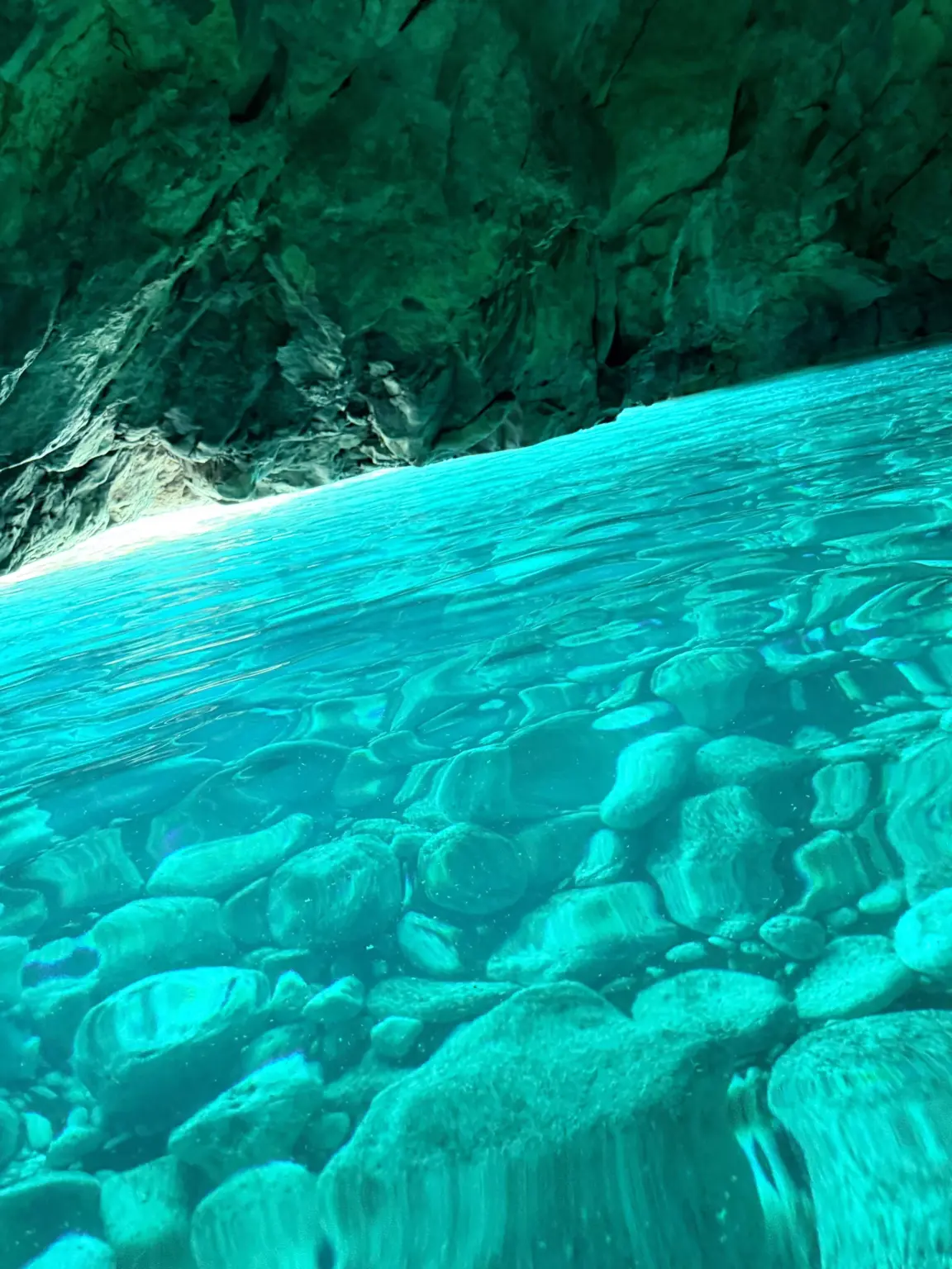 Clear turquoise water over rounded pebbles inside the Blue Cave, below a dark green rock wall and a bright band of light