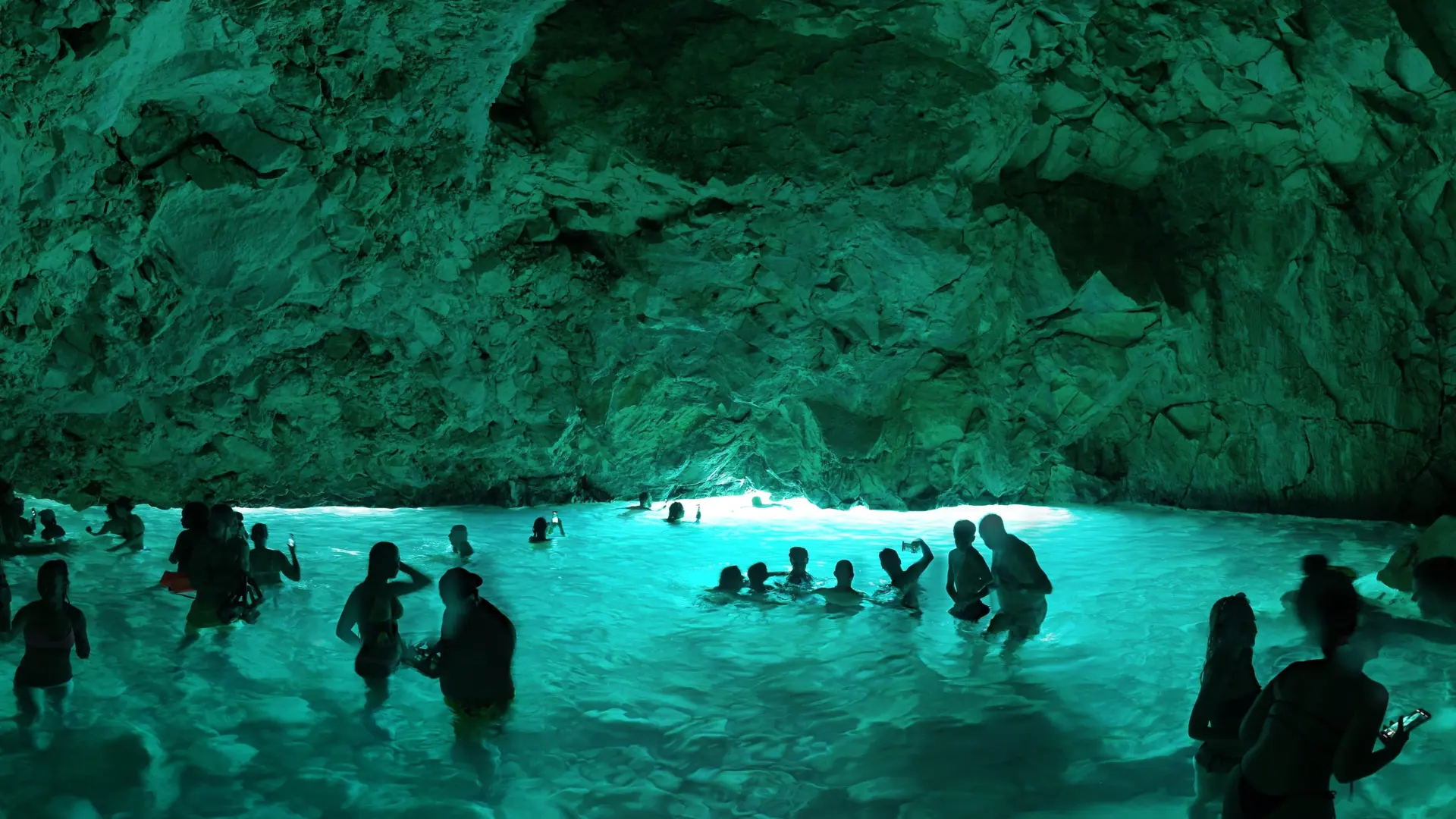 Visitors in turquoise water under the low rock roof of the Blue Cave, with daylight at the far end