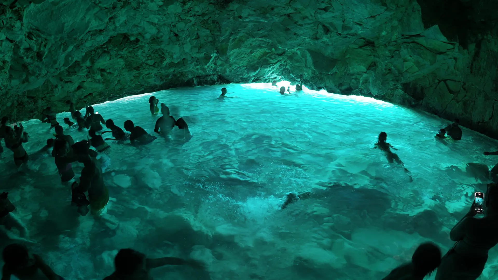 Swimmers in turquoise water inside a low sea-cave chamber, with daylight showing at the far end